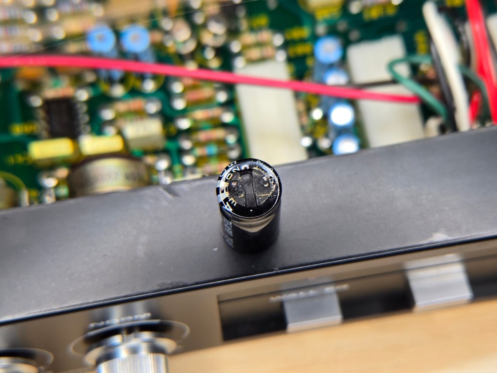 A view of the bottom side of the removed capacitor from the previous image, showing that the plastic sheath is crinkled slightly - evidence of leakage producing heat - and there is a crust of leaked fluid around the edges.