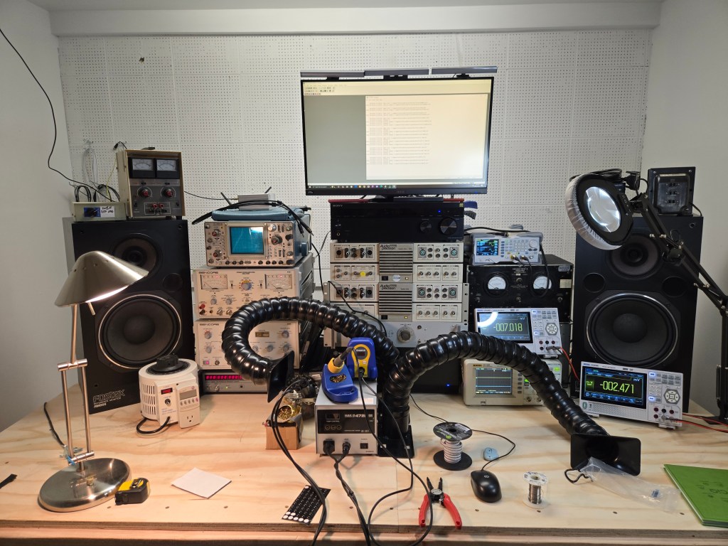 A photo of the new workbench showing two large studio monitors on either side, with multiple stacks of test equipment in the center showing various analog and digital displays.