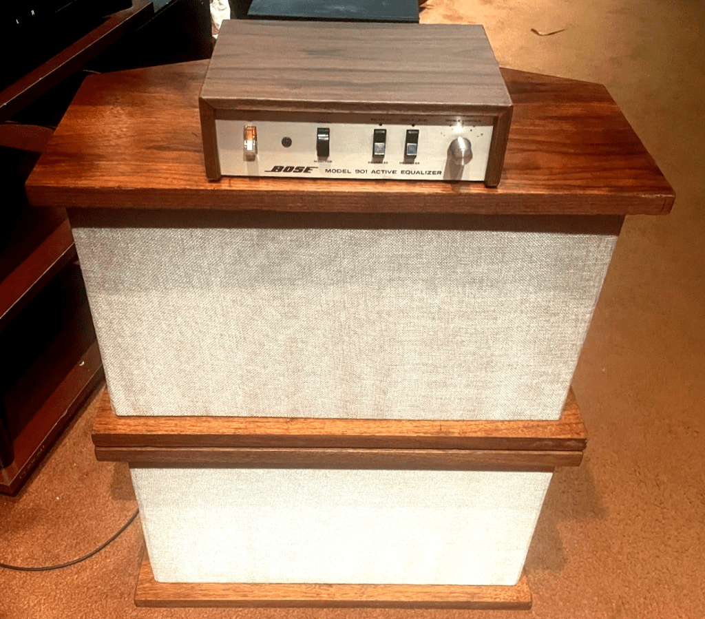 A pair of Bose 901 Series I speakers stacked togther with the equalizer on top. The equalizer is turned on and has a glowing power switch; the speakers are facing in the correct orientation with the flat side carrying the single driver facing towards the listener.