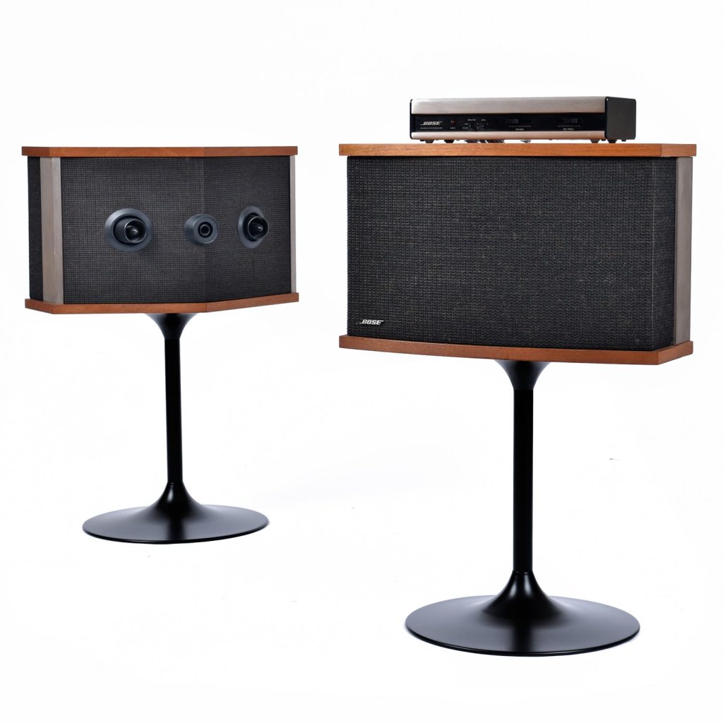 Promotional photo showing the Bose 901 Series V speakers, front and rear, on black tulip stands with their Active Equalizer sitting on top.